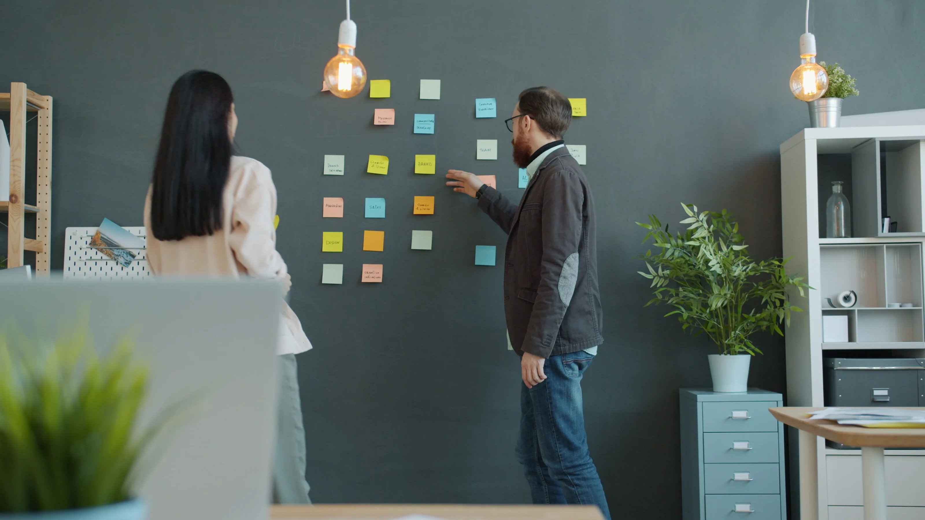 Dos personas frente a una pared con notas de trabajo, ordenando las prioridades de un proyecto